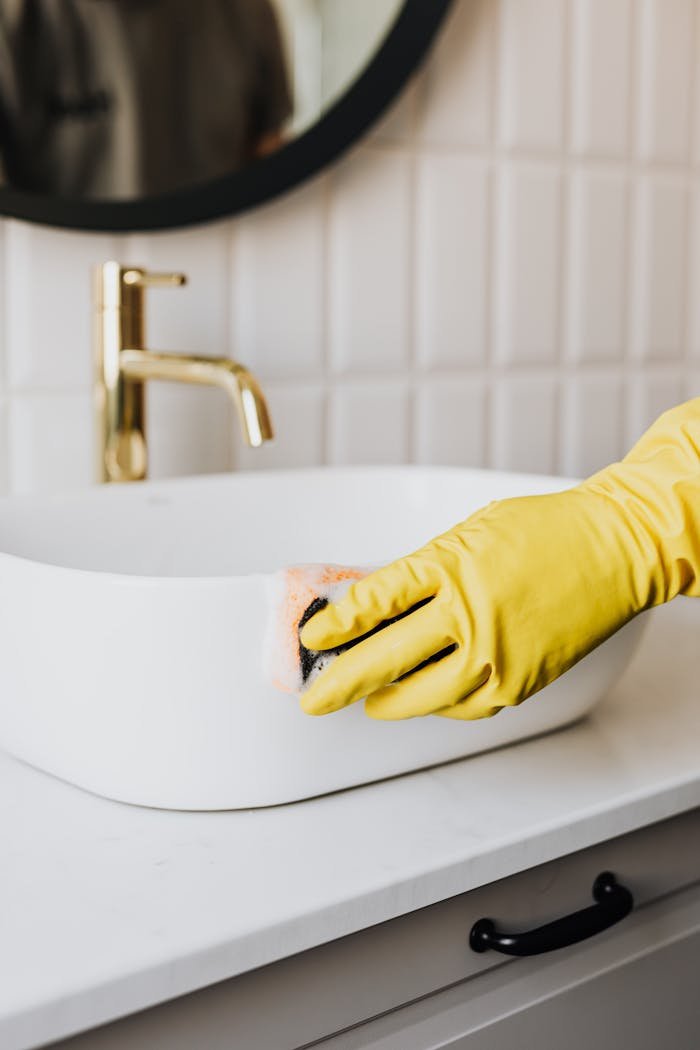 gallery-1 Crop faceless person in yellow rubber protective glove wiping surface of white sink in bathroom with sponge and cleaning agent