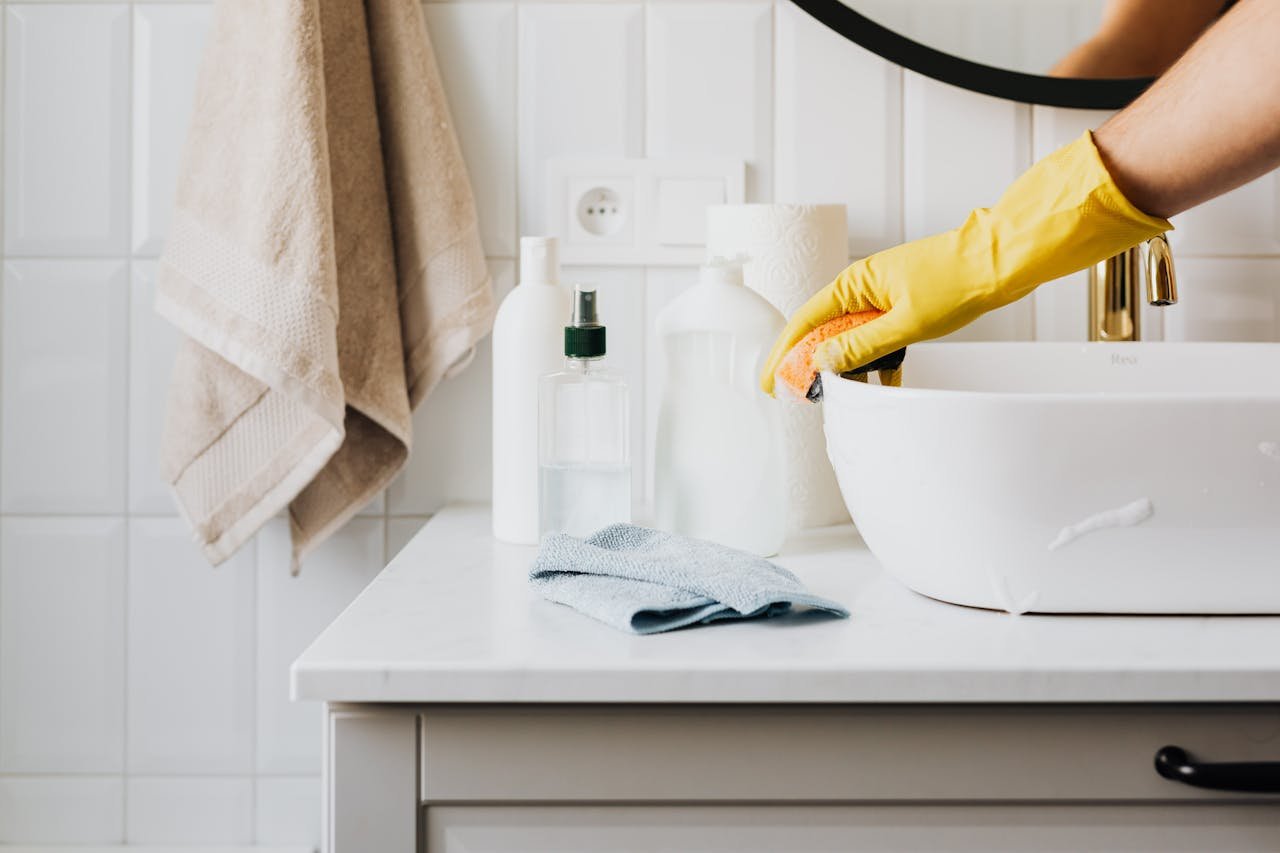 about-01 Person cleaning a bathroom sink with yellow gloves and cleaning products on a countertop.