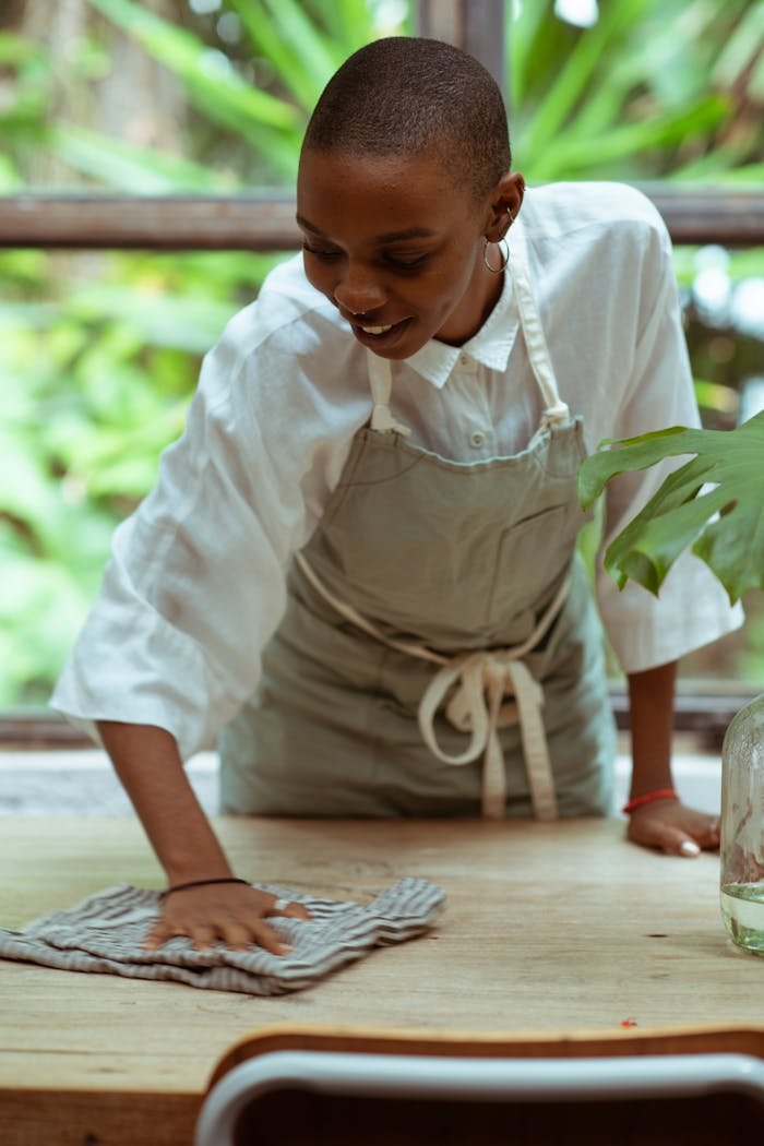 gallery-2 Positive smiling black woman with short haircut wearing apron cleaning wooden table by using microfiber cloth while standing against blurred big window and green lush park