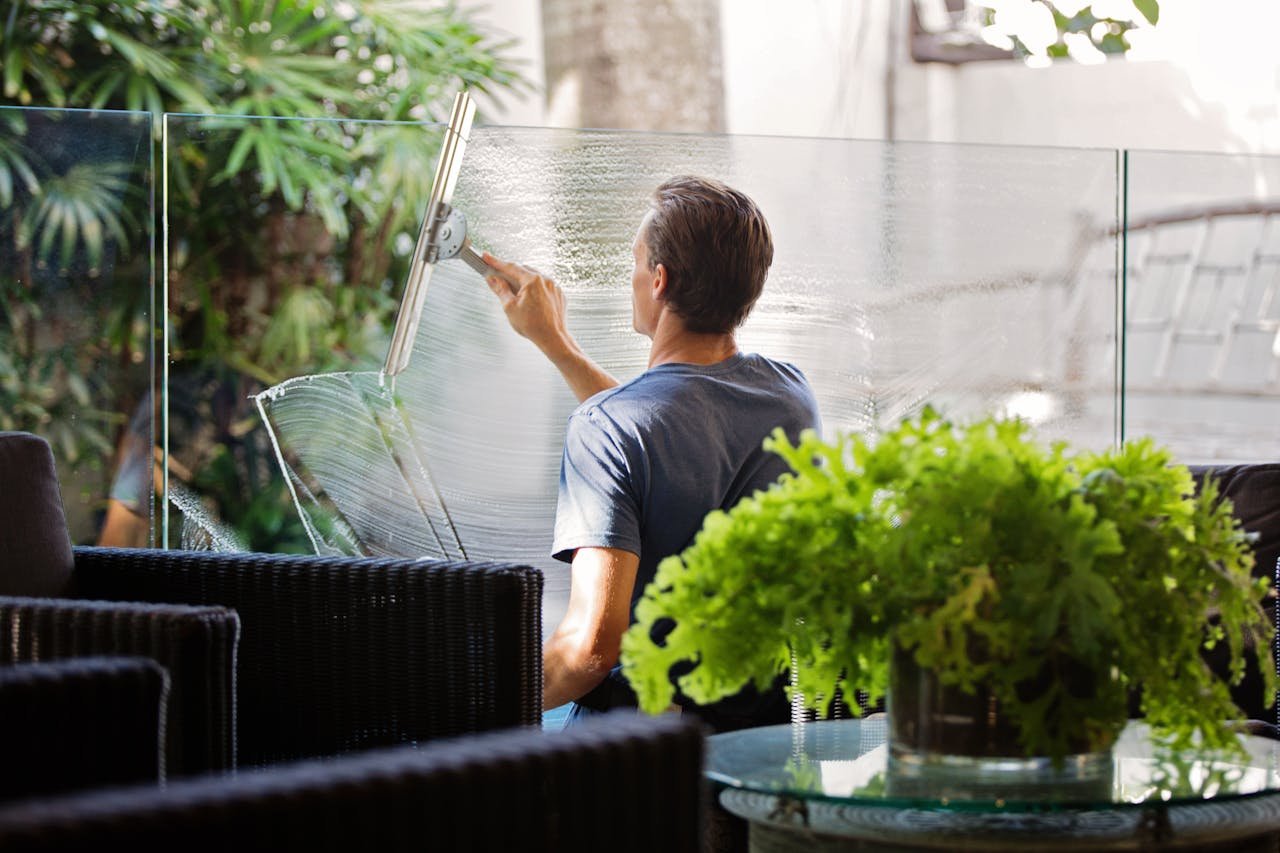 hero-img-02 A man cleaning a glass barrier outdoors with a squeegee, surrounded by greenery.